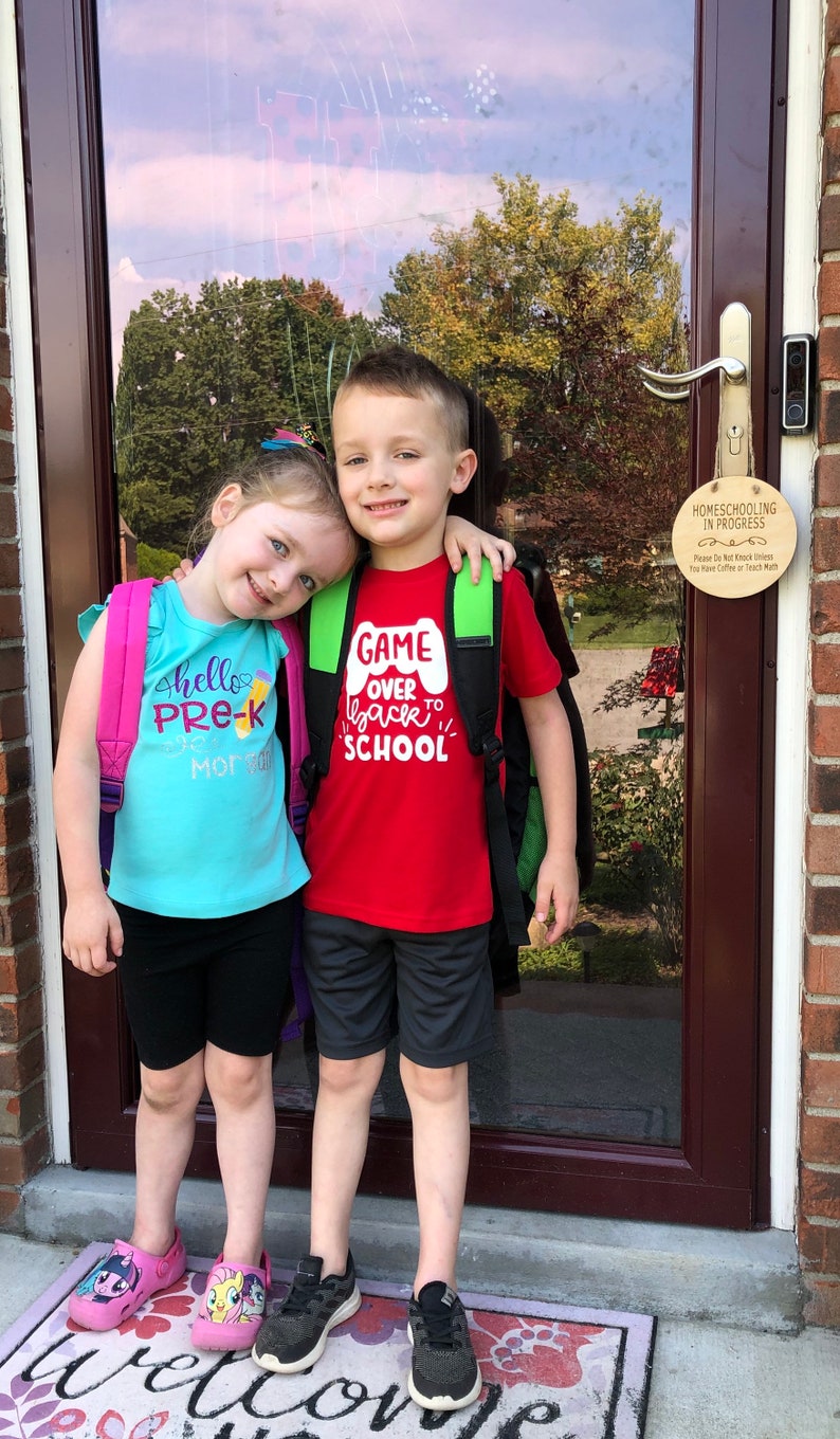 May include: Two children, a girl and a boy, stand in front of a door with a sign that reads "Homeschooling in Progress". The girl is wearing a turquoise shirt with the text "hello pre-k" and a pink backpack. The boy is wearing a red shirt with the text "Game Over Back to School" and a green backpack. They are both smiling.