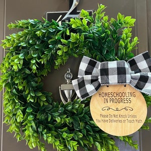 May include: A wooden sign with the text "Homeschooling in Progress" and a black and white checkered bow hanging on a green wreath. The sign also says "Please Do Not Knock Unless You Have Deliveries or Teach Math."