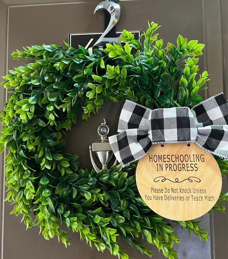 May include: A wooden sign with the text "Homeschooling in Progress" and a black and white checkered bow hanging on a green wreath. The sign also says "Please Do Not Knock Unless You Have Deliveries or Teach Math."