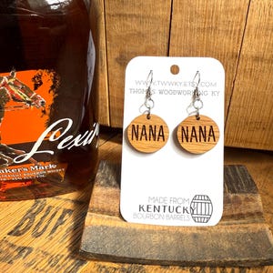 May include: A pair of wooden earrings with the word "NANA" engraved on each. The earrings are made from repurposed Kentucky bourbon barrels. The earrings are displayed on a white card with the website address and the text "Made from Kentucky Bourbon Barrels".