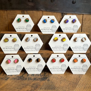 May include: Assortment of 10 pairs of earrings made from repurposed Kentucky bourbon barrels. Each pair features a different color combination of wood and paint. The earrings are displayed on white hexagonal cards with the website address and text "Made from Kentucky Bourbon Barrels".
