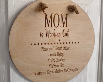 May include: A round wooden sign with the text "MOM is Working Out" and a list of reasons not to disturb: "You're Dying", "You're Bleeding", "You're on Fire", and "The Amazon Guy is Matthew McConaughey".