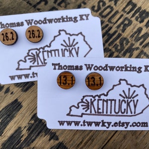 May include: Wooden stud earrings with the numbers "26.2" and "13.1" engraved on them. The earrings are displayed on white cards with the text "Thomas Woodworking KY" and a Kentucky outline. The background is a wooden surface.