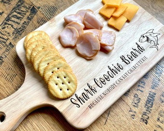 May include: A wooden cutting board with a shark design and the text "Shark Coochie Board because nobody can say charcuterie". The board has a handle and is decorated with crackers, cheese, and sliced meat.