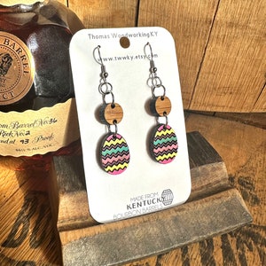 May include: A pair of colorful, wooden earrings with a zig-zag pattern. The earrings are made from repurposed Kentucky bourbon barrels and feature a silver hook. The card reads "Made from Kentucky Bourbon Barrels" and "Thomas Woodworking KY www.twwky.etsy.com".