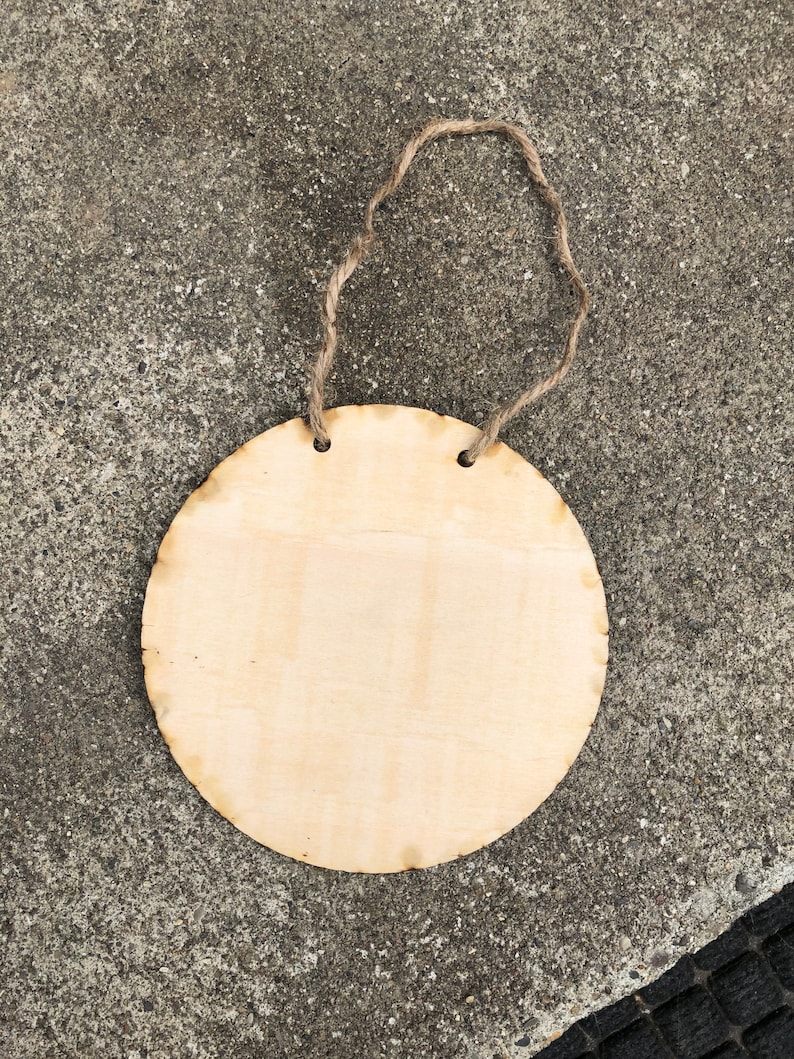 May include: A round, unfinished wooden sign with a rope hanger. The sign is blank and ready to be decorated.