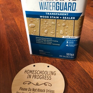May include: A can of Olympic Waterguard Transparent Wood Stain + Sealer in Maple Brown. The can is labeled with the product name, color, and protection details. It states that it protects up to 3 years on decks and 4 years on fences and siding. The can is sitting on a wooden surface next to a round wooden sign that says "Homeschooling in Progress" with a decorative swirl and the text "Please Do Not Knock Unless You Have Coffee or Teach Math".