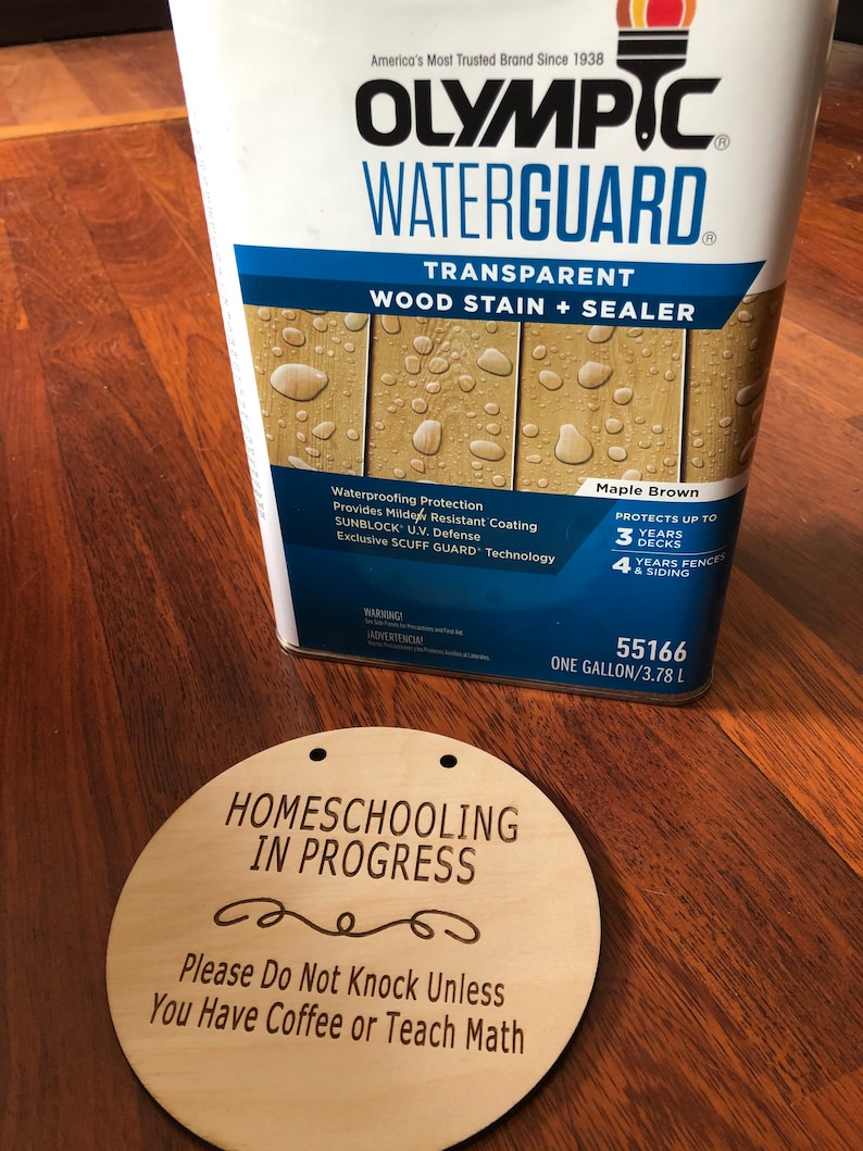 May include: A can of Olympic Waterguard Transparent Wood Stain + Sealer in Maple Brown. The can is labeled with the product name, color, and protection details. It states that it protects up to 3 years on decks and 4 years on fences and siding. The can is sitting on a wooden surface next to a round wooden sign that says "Homeschooling in Progress" with a decorative swirl and the text "Please Do Not Knock Unless You Have Coffee or Teach Math".