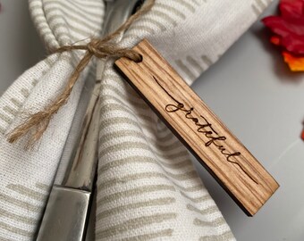 May include: A wooden place card with the word "grateful" engraved on it. The place card is tied to a fork and napkin with twine.