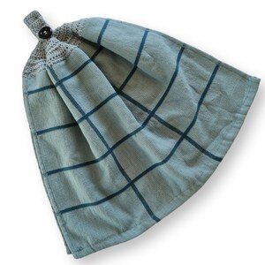 May include: A hanging kitchen towel in a muted teal color with a grid pattern in a darker teal. The top of the towel has a crocheted section in a light gray color, with a dark brown button for hanging. The towel is made of a soft, absorbent material.