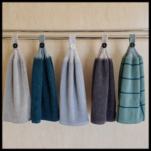 May include: Five hanging hand towels in shades of grey, teal, and blue. Each towel has a crocheted top with a button closure. The towels are displayed on a silver bar against a neutral tiled background.