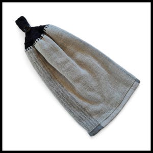 May include: A grey and black hanging kitchen towel. The top section is black with a button fastening. The towel is a light grey colour with a darker grey border and accents. The towel is made of a textured fabric.
