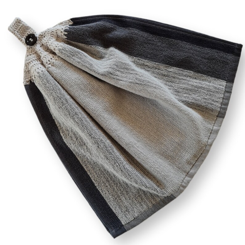 May include: A hanging kitchen towel with a button closure. The towel features a color block design with sections of gray, beige, and dark gray. The top portion is crocheted with a button for hanging. The towel is made of a textured fabric.