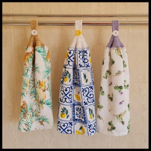 May include: Three hanging kitchen towels with crochet tops and button fastenings. The towels display vibrant patterns: a parrot and tropical leaves, blue and white tile design with lemons, and hummingbirds with leaves.