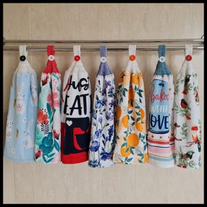 May include: Seven hanging kitchen towels with crochet tops and button fastenings. Designs include floral, bunny, and fruit patterns, along with the phrases "Just Eat" and "Baked with Love". The towels are displayed on a metal rail.