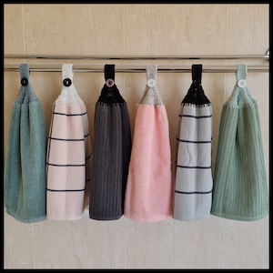 May include: Six crocheted tea towels hanging on a rack. The towels are in various colours, including green, pink, grey, and white. Each towel has a black crocheted loop at the top and a white button.