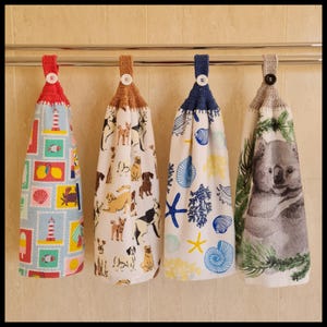 May include: Four hanging kitchen towels with crochet tops and button fastenings. The towels display diverse patterns: a nautical theme with stamps, a dog print, a coastal design with seashells, and a koala bear with greenery.