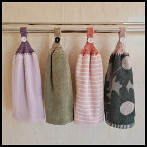 May include: Four hanging kitchen towels with crochet tops and button fastenings. The towels are in various colours and patterns: lavender, olive green, pink stripes, and a dark green floral design. They hang from a silver bar.