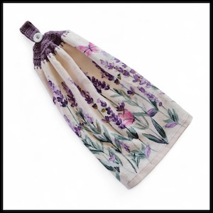 May include: A hanging kitchen towel with a lavender floral design. The towel has a cream background with purple lavender sprigs, pink flowers, and green leaves. A purple crocheted top with a white button completes the design.