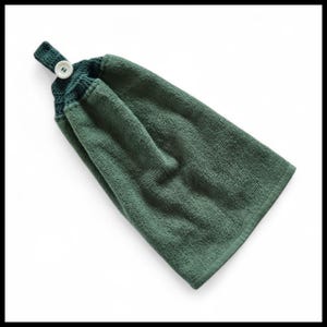 May include: A sage green terry cloth kitchen towel featuring a dark forest green crocheted top section with a single white button closure. The towel is folded and hangs from a small loop at the top, showcasing a soft, absorbent fabric texture.