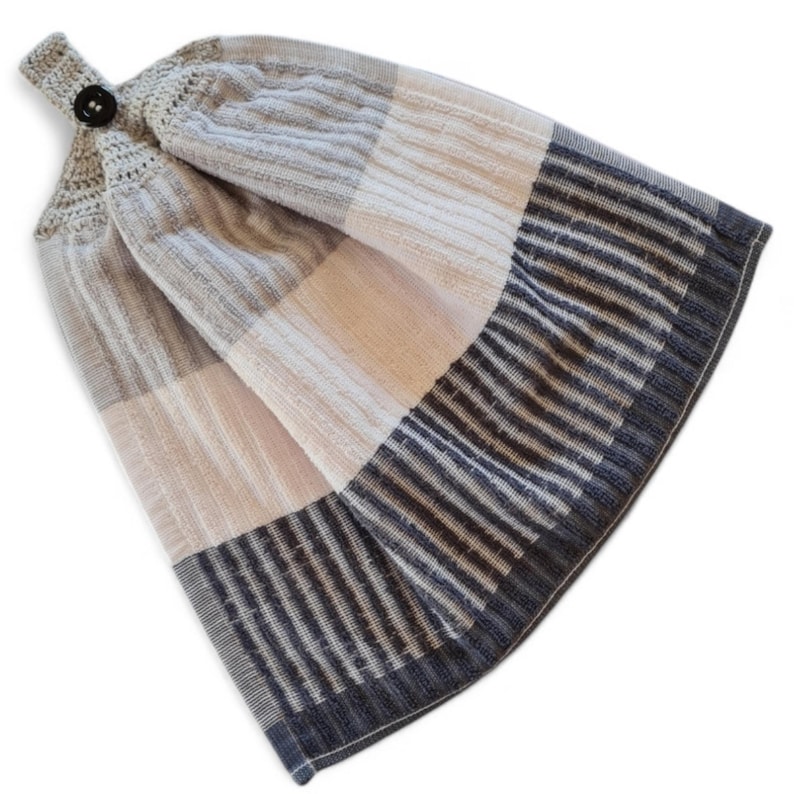 May include: A hanging kitchen towel with a button closure. The towel features a patchwork design with gray, off-white, and navy blue stripes. The top portion is a solid gray color.