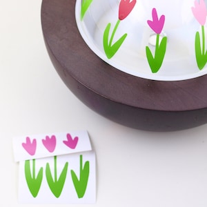 May include: A wooden base with a glass dome featuring a design of pink and green tulips.  The dome is removable and there are extra tulip stickers on a white sheet.