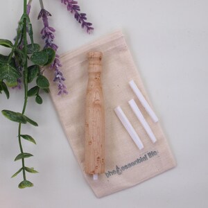 May include: A light-colored wooden essential oil diffuser with a matching canvas bag and three white diffuser sticks. The bag has the text "the essential life" printed on it. Lavender and greenery are in the background.