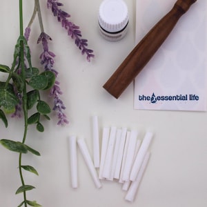 May include: An overhead shot of essential oil diffuser supplies. The image includes a small white bottle, a dark wooden tool, white diffuser sticks, and a card with the text "the essential life". Lavender sprigs add a touch of color.