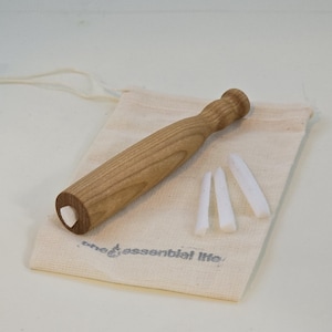 May include: A light brown wooden massage tool with a rounded handle and a white insert at the base. The tool rests on a beige canvas bag with the text "one essential life" printed on it, along with three white, triangular massage sticks.