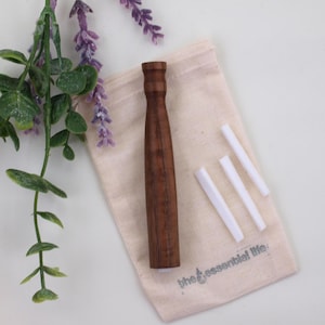 May include: A dark brown wooden tool with a rounded top and a cylindrical body, resting on a beige pouch with the text "the essential life". Three white rectangular objects are next to the pouch. Purple and green foliage is in the background.