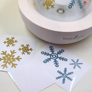 May include: Three sheets of white paper with gold and blue snowflake stickers. The stickers are ready to be applied to a white diffuser with the words "Young Living Essential Oils" printed on it.