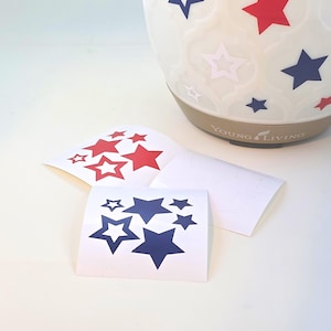 May include: White essential oil diffuser with red and blue star decals. Two sheets of star-shaped decals in red and blue are in the foreground. The diffuser has the words "Young Living" printed on it.