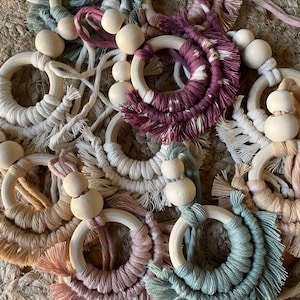 May include: A collection of macrame rings in various colors, including beige, pink, and teal. Each ring features a wooden bead and a fringe detail. The rings are arranged on a woven surface.