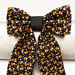 Puede incluir: Un lazo decorativo grande con un centro negro y un patrón de caramelos de maíz. El lazo presenta una pieza central negra y colas largas y puntiagudas. El patrón de caramelos de maíz incluye acentos naranjas, amarillos, blancos y morados sobre un fondo negro.