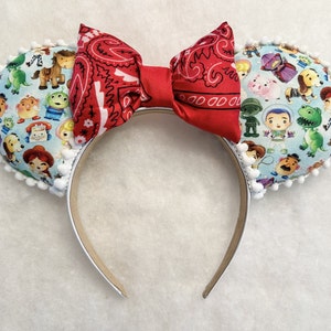 May include: Toy Story themed Minnie Mouse ears with a red bandana bow and white pom pom trim. The ears are covered in a blue fabric with a pattern of Toy Story characters.
