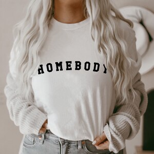 May include: White long-sleeve shirt with the text "HOMEBODY" printed in black.