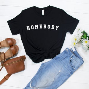 May include: Black t-shirt with white text that reads "HOMEBODY". The shirt is styled with blue jeans, a brown fanny pack, and brown sandals.
