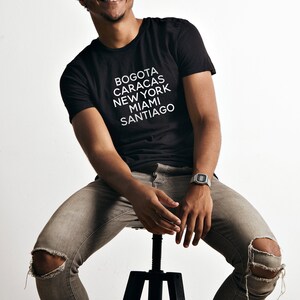 May include: A man wearing a black t-shirt with the words "BOGOTA CARACAS NEW YORK MIAMI SANTIAGO" printed in white. He is wearing grey jeans with holes in the knees and a silver watch on his left wrist.