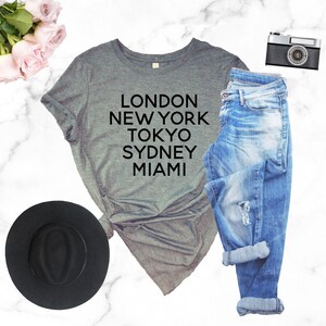 May include: Gray t-shirt with black text that reads "LONDON NEW YORK TOKYO SYDNEY MIAMI". The shirt is paired with blue jeans, a black hat, and a vintage camera.