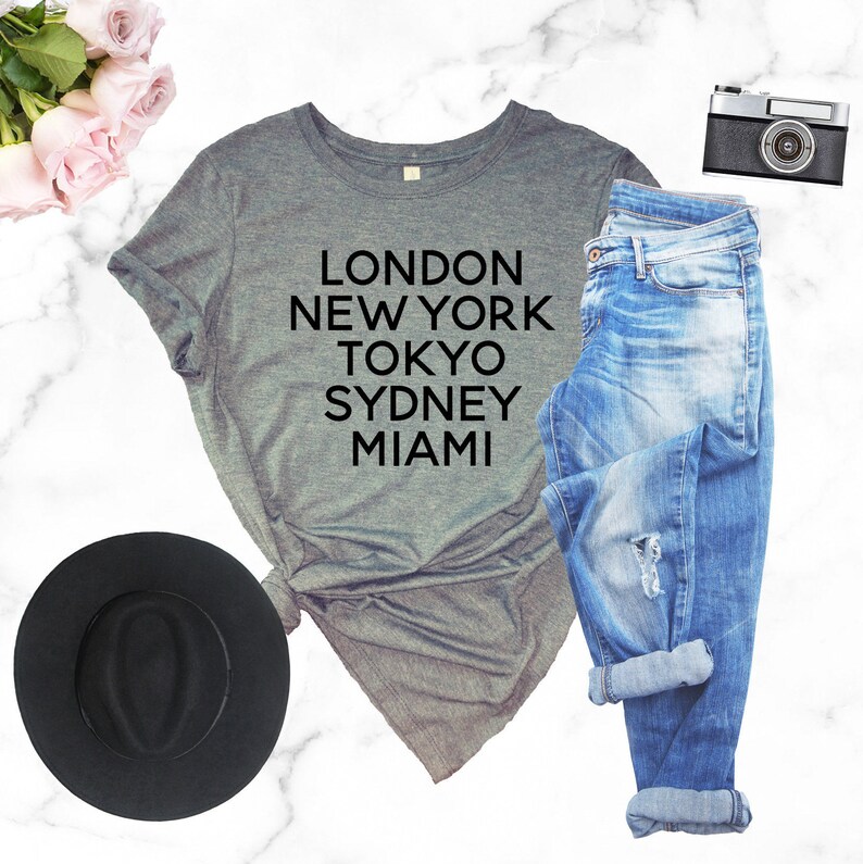 May include: Gray t-shirt with black text that reads "LONDON NEW YORK TOKYO SYDNEY MIAMI". The shirt is paired with blue jeans, a black hat, and a vintage camera.