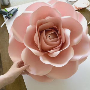 May include: A large, handmade paper flower in a soft blush pink. The flower has multiple layers of petals, creating a dimensional effect. The center of the flower is tightly wound, and the outer petals are gently curved. This decorative paper flower is ideal for event decor.