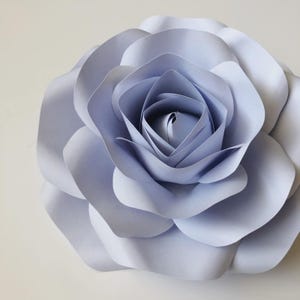 Op de afbeelding: Een grote, handgemaakte papieren bloem in een zachte periwinkeltint. De bloemblaadjes zijn gelaagd, waardoor een realistische roosvorm ontstaat. De bloem is een decoratief item, perfect voor wanddecoratie of evenementstyling.