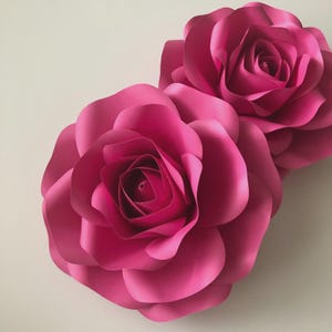 Op de afbeelding: Twee grote, levendige roze papieren rozen. De gelaagde bloemblaadjes creëren een realistisch bloemdesign. De rozen staan tegen een effen achtergrond, waardoor hun kleur en vorm worden benadrukt. Deze decoratieve bloemen zijn ideaal voor evenementen of interieurdecoratie.