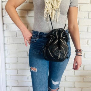 Può includere: Borsa nera con coulisse, borchie color oro e un design geometrico. La borsa è indossata a tracolla. La borsa è in pelle e ha una chiusura con coulisse. La borsa è alta circa 20 cm e larga 18 cm.