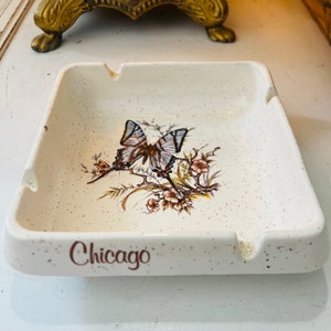 Puede incluir: Cenicero de cerámica blanca con un acabado moteado. Presenta una mariposa marrón con un patrón blanco y gris sobre un diseño floral. La palabra "Chicago" está impresa en marrón en la parte inferior.