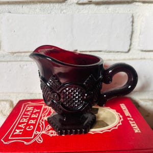 May include: A ruby red glass creamer with intricate embossed detailing. The creamer features a spout, a handle, and a decorative pattern around the body. It sits on a red book with the title "Marian Grey" visible. The creamer is a vintage piece.