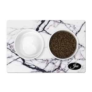 May include: White marble pet food mat with two white ceramic bowls, one filled with brown kibble. The mat has a black oval with the name "Jax" written in white.