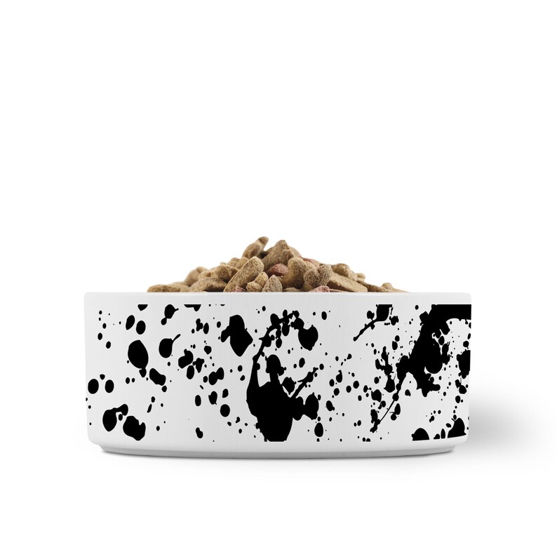 Puede incluir: Taz&oacute;n de cer&aacute;mica blanco para mascotas con un dise&ntilde;o de pintura salpicada negra. El taz&oacute;n est&aacute; lleno de comida seca para mascotas.