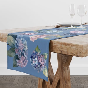 May include: A blue table runner with a watercolor floral design featuring hydrangeas. The runner has a light blue background with pink and blue hydrangeas and green leaves.