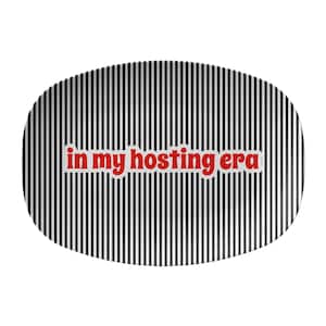 May include: An oval-shaped serving platter with a black and white striped background. The words "in my hosting era" are printed in red, outlined in white, across the center of the platter. The platter is suitable for serving food or as a decorative piece.